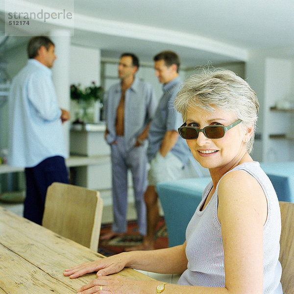 Reife Frau sitzt am Tisch  drei Männer stehen im Hintergrund