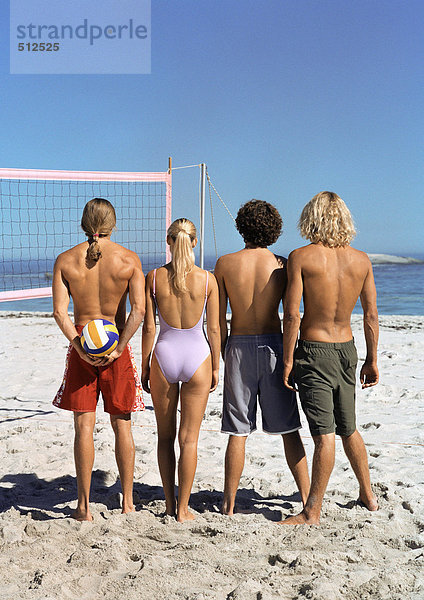 Vier junge Leute stehen am Strand mit Volleyball  Rückansicht.