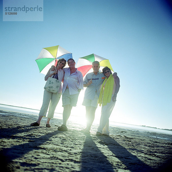 Zwei reife Paare stehen am Strand und halten Sonnenschirme.