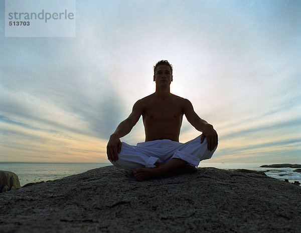 Mann im indischen Stil auf Felsen sitzend  hinterleuchtet  volle Länge  Himmel und Meer im Hintergrund