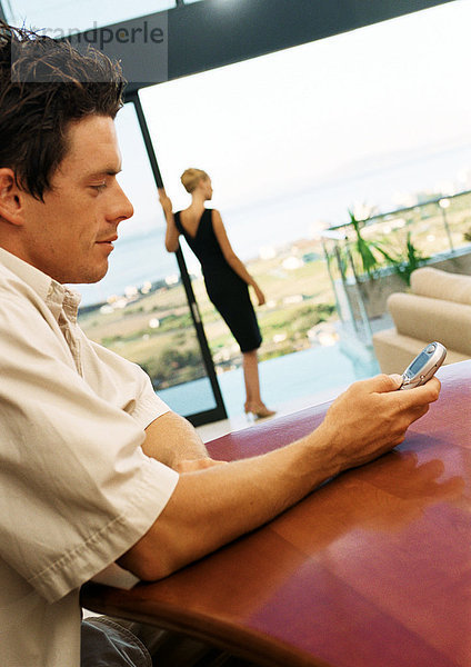 Mann mit Handy  Frau auf der Terrasse im Hintergrund