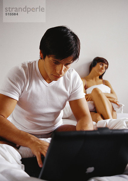 Mann arbeitet am Laptop im Bett  Frau im Hintergrund.