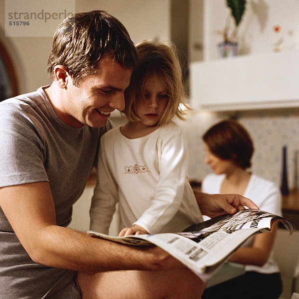 Vater sitzend mit Tochter auf Papier schauend  Frau im Hintergrund