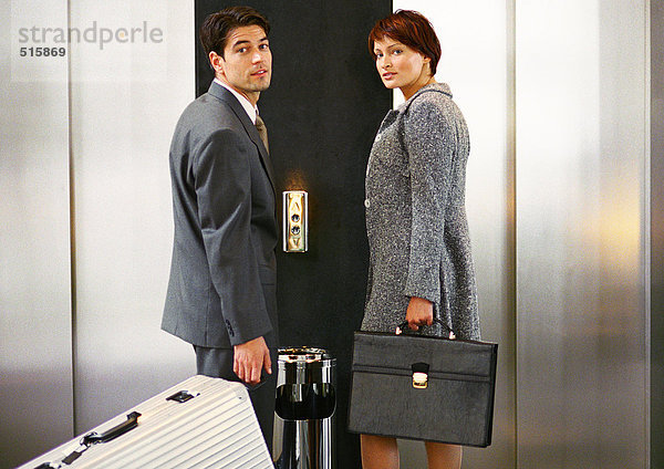 Geschäftsmann und Geschäftsfrau warten auf den Aufzug und schauen über die Schultern in die Kamera.
