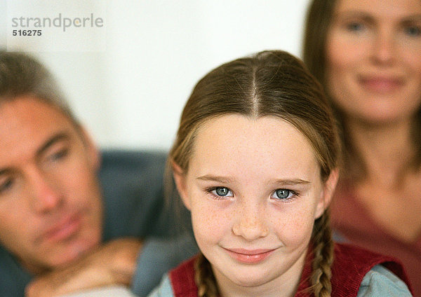 Kleines Mädchen mit lächelnden Zöpfen  erwachsener Mann und Frau im Hintergrund  Nahaufnahme  Porträt