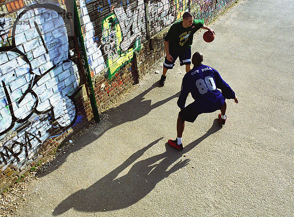 Zwei Männer spielen Basketball vor einer graffitierten Wand auf einem städtischen Spielplatz.