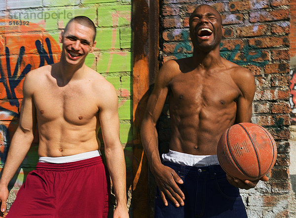 Zwei Männer ohne Hemd stehen und lachen neben der graffitierten Wand  einer hält Basketball.