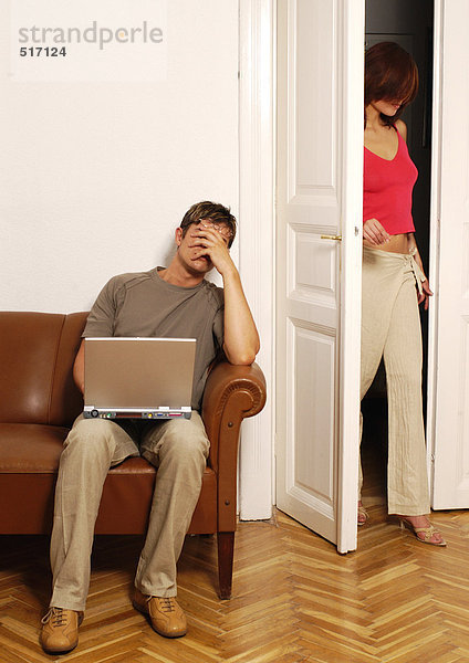 Junger Mann sitzt mit Laptop  reibt die Augen  während die Frau ins Zimmer kommt.
