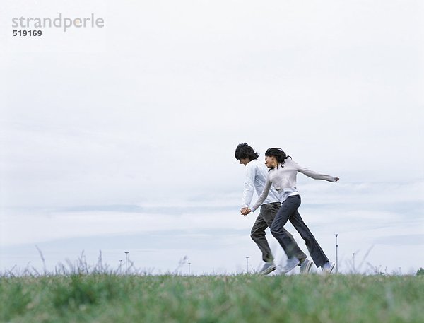 Junger Mann und junge Frau halten sich an den Händen und laufen auf Gras.