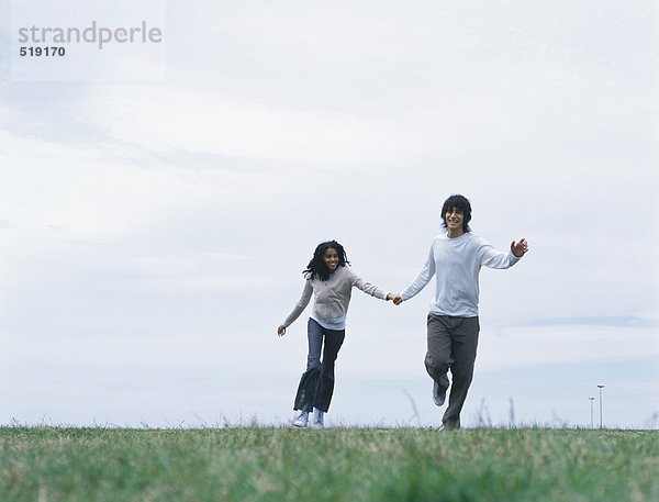 Junger Mann und junge Frau halten sich an den Händen und laufen auf Gras.