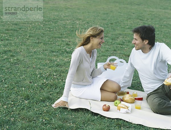 Junger Mann und junge Frau sitzen auf einer Decke auf Gras und machen ein Picknick.