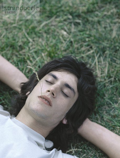 Junger Mann auf Gras liegend mit geschlossenen Augen und einem Stück Gras im Mund.
