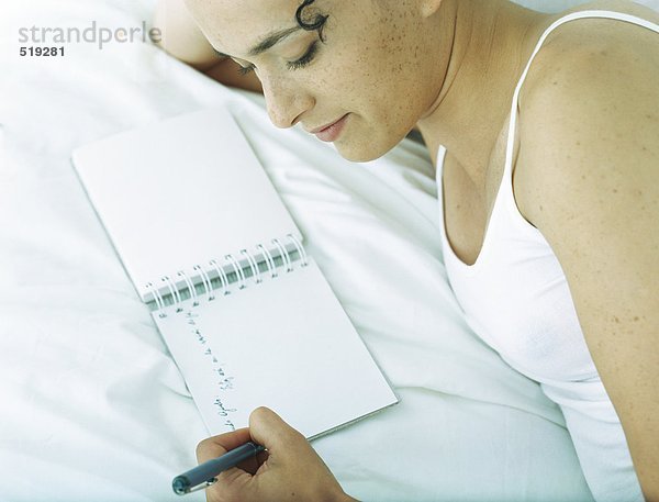 Frau liegt seitlich auf dem Bett und schreibt in Notizbuch mit Stift