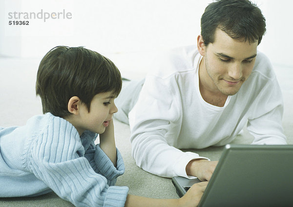 Mann und kleiner Junge liegen auf dem Boden und arbeiten am Laptop.