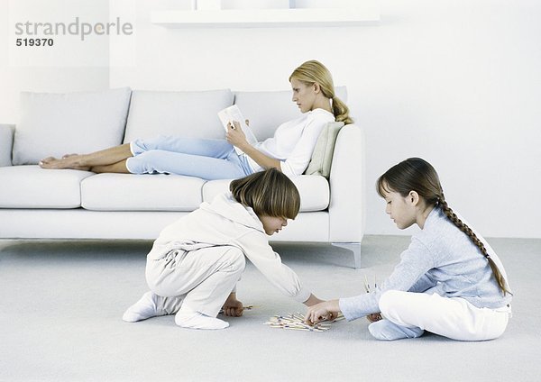 Junge und Mädchen sitzen auf dem Boden  Frau sitzt auf dem Sofa und liest im Hintergrund.