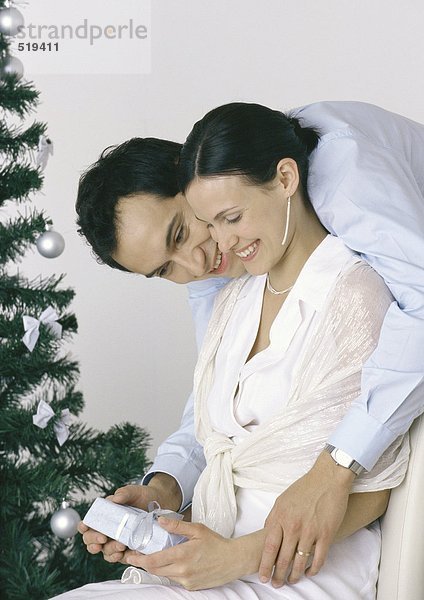 Frau hält Geschenk  Mann beugt sich über die Schulter  Weihnachtsbaum im Hintergrund