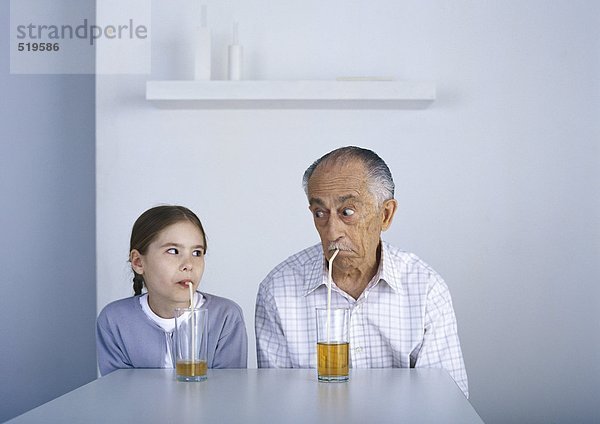 Großvater und Enkelin sitzen am Tisch  trinken mit Strohhalmen und schauen sich an.