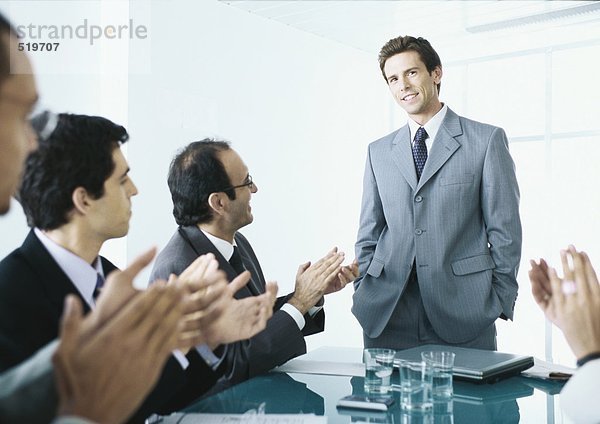 Sitzende Geschäftsleute applaudieren Geschäftsmann am Ende des Tisches stehend