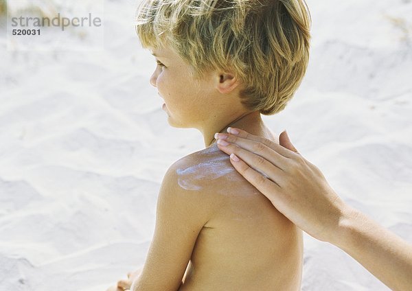 Kleiner Junge am Strand mit Sonnencreme von Mutter in die Schulter gerieben