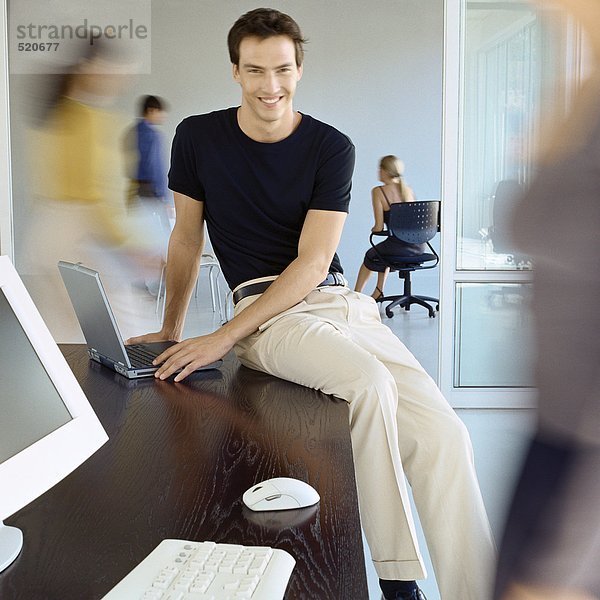 Mann sitzt auf Schreibtischkante im geschäftigen Büro