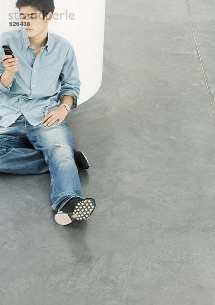 Junger Mann sitzt auf dem Boden und schaut auf das Handy