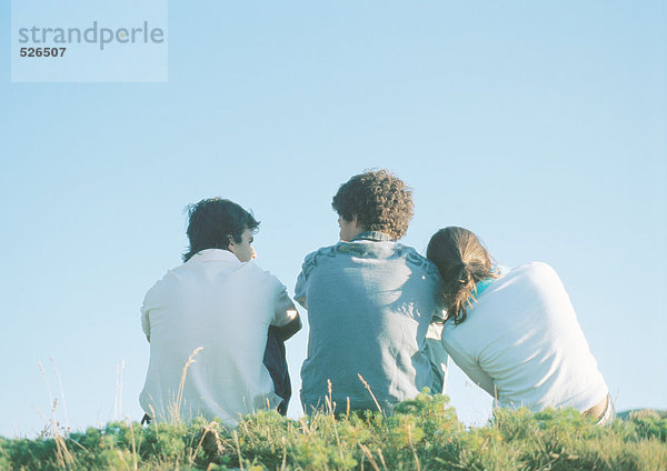 Drei Teenager sitzen auf Gras  ein Mädchen lehnt den Kopf auf die Schulter des Jungen.