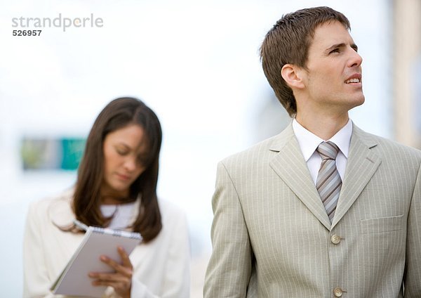 Geschäftsmann schaut auf  während die Assistentin im Hintergrund Notizen macht.