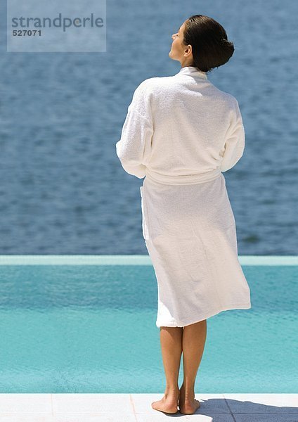 Frau im Bademantel am Pool mit Blick aufs Meer  Rückansicht