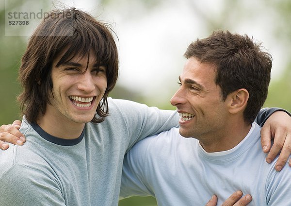 Zwei junge männliche Freunde lächeln mit den Armen um die Schultern.