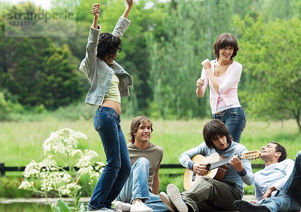 Freundeskreis im Freien  junge Frauen tanzen  junger Mann spielt Gitarre