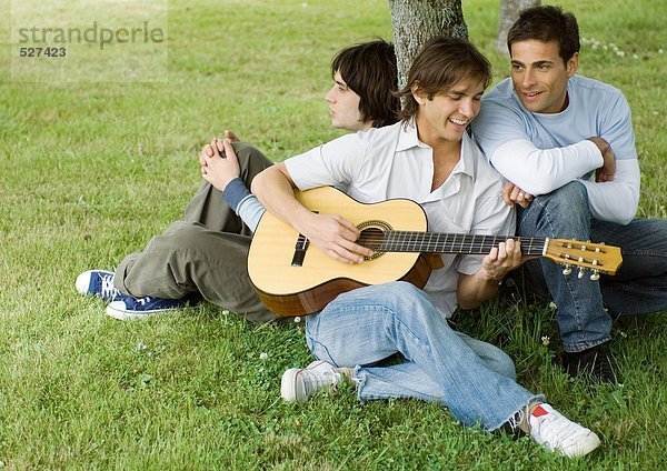 Junge männliche Freunde sitzen neben einem Baum und spielen Gitarre.