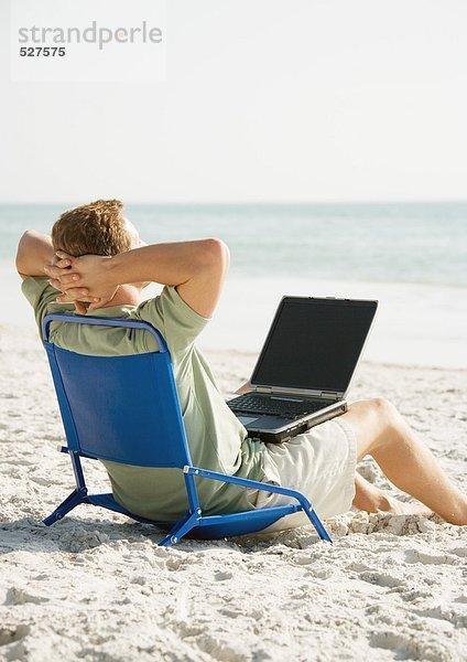 Mann am Strand sitzend mit Computer auf dem Schoß