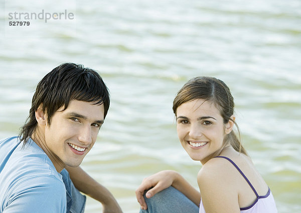 Junges Paar sitzt neben dem Wasser  lächelt und schaut in die Kamera.