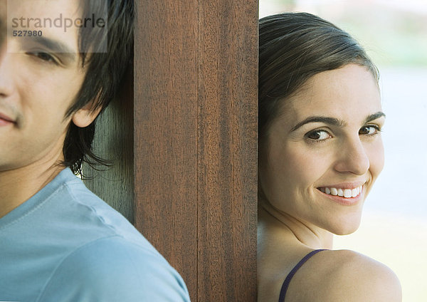 Junges Paar steht Rücken an Rücken mit einem Holzpfosten dazwischen  lächelnd  Portrait  Teilansicht