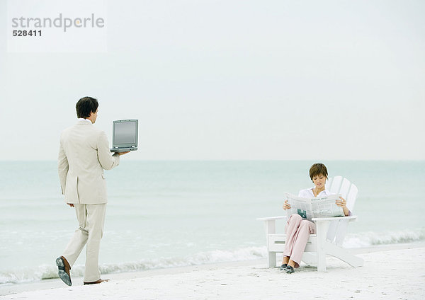 Frau sitzt am Strand und liest Zeitung als Geschäftsmann mit offenem Laptop