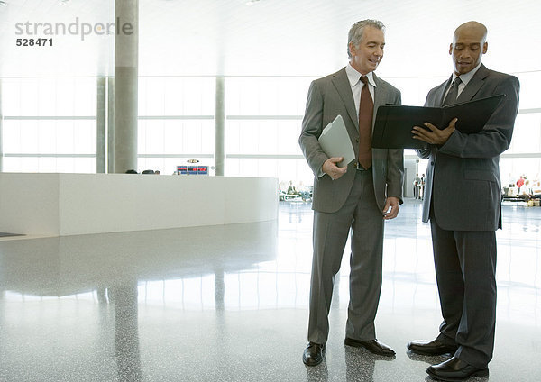 Zwei Geschäftsleute stehen in der Flughafenhalle und schauen sich die Tagesordnung an.