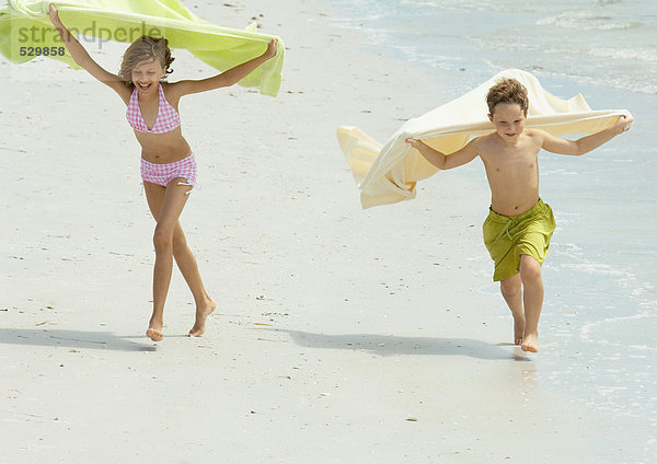 Zwei Kinder laufen am Strand und halten Handtücher im Wind.