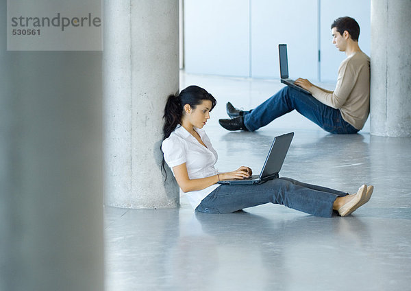 Junge Frau und junger Mann sitzen auf dem Boden und benutzen Laptops.