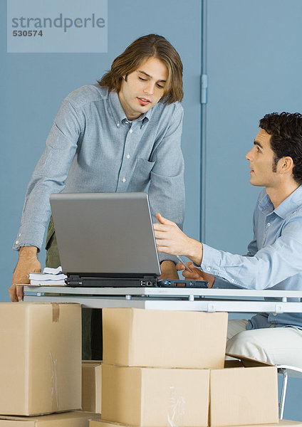 Geschäftsleute mit Laptop  die an der Tischplatte sitzen und von Kartons gestützt werden.