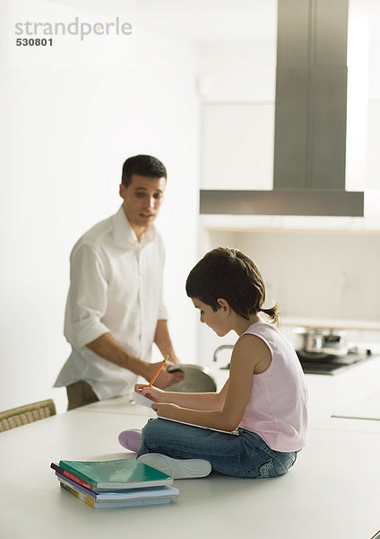 Kind sitzt auf der Theke und macht Hausaufgaben  während Vater kocht.