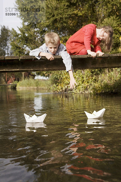 Junge (10-12) und Mädchen (7-9) auf der Brücke beobachten Papierboote im Wasser