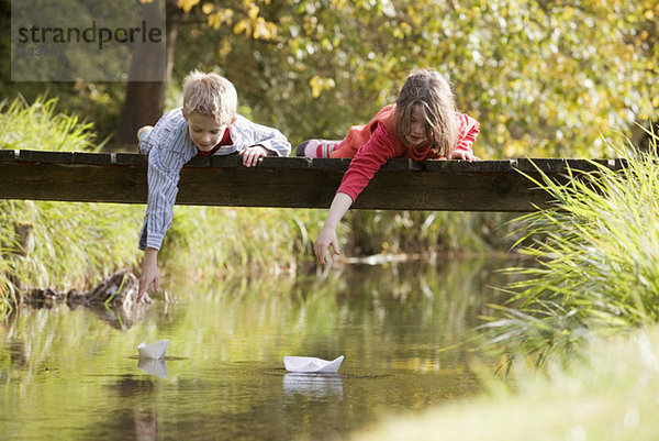 Junge (10-13) und Mädchen (7-9) liegen auf der Brücke und beobachten Papierboote im Wasser.