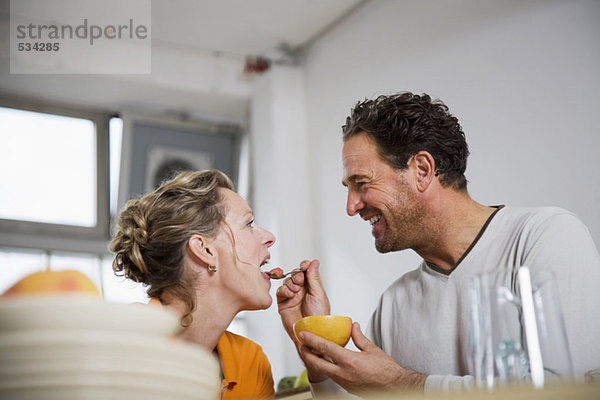Reifer Mann füttert Grapefruit an Frau in der Küche  lächelnd  Blickwinkel niedrig