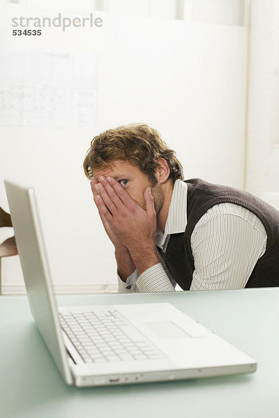 Young man sitting by laptop  face covered with hands