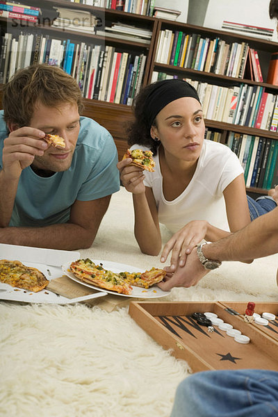 Drei junge Leute liegen auf dem Boden und essen Pizza.