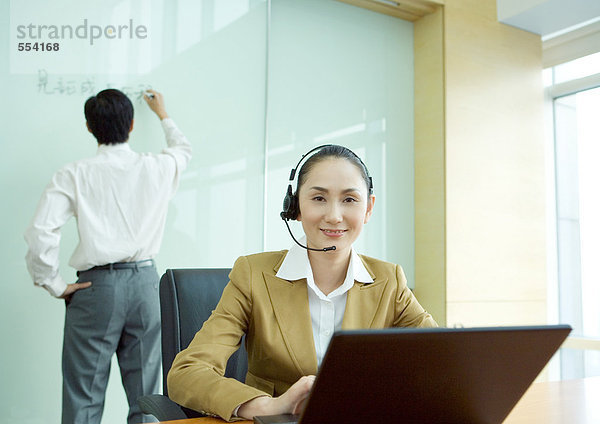 Geschäftsfrau mit Headset und Laptop  Kollegin schreibt auf Whiteboard im Hintergrund