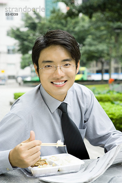 Geschäftsmann beim Mittagessen mit Essstäbchen und Zeitung lesen  lächelnd vor der Kamera