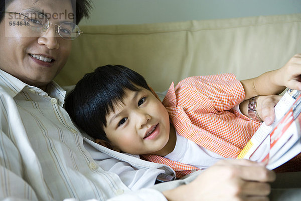 Vater und Sohn sitzen auf dem Sofa mit Buch und lächeln in die Kamera.