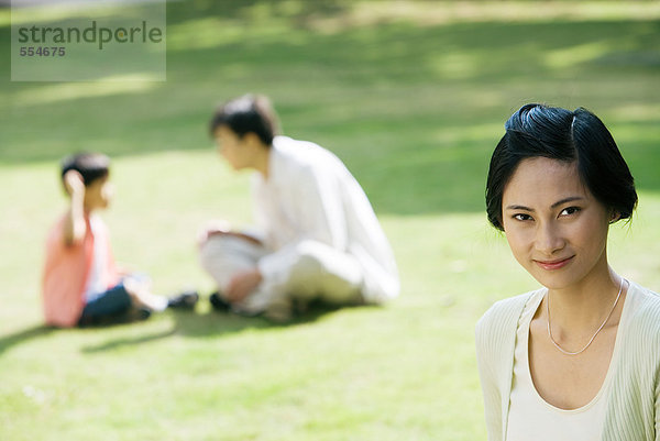 Frau lächelt in die Kamera  Mann und Sohn sitzen auf Gras im Hintergrund