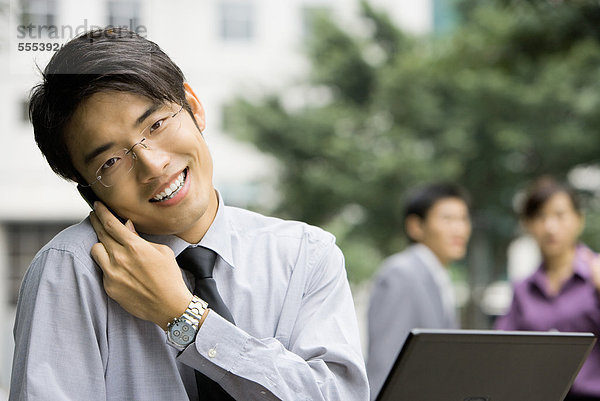 Junger Geschäftsmann hält Handy gegen die Schulter  lächelt in die Kamera